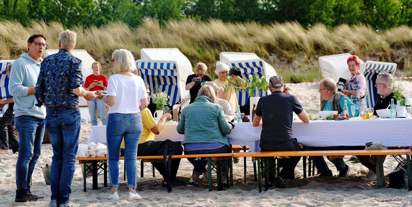Mehrere Menschen sitzen an einem langen Tisch am Strand und unterhalten sich.