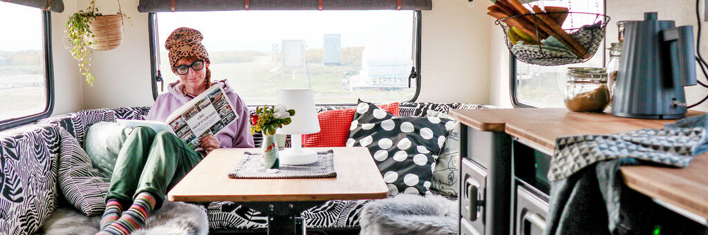 Eine Person liest in einem gemütlichen Wohnmobil mit stilvollem Interieur. Auf dem Tisch stehen Blumen und ein Glas. Das Wohnmobil bietet einen Blick auf eine grüne Landschaft.