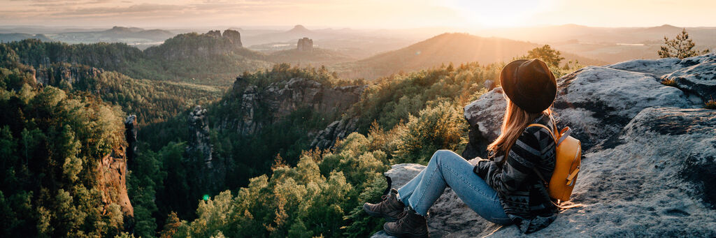 Frau sitzt auf einem Felsen und genießt den Sonnenuntergang über einer bewaldeten Landschaft
