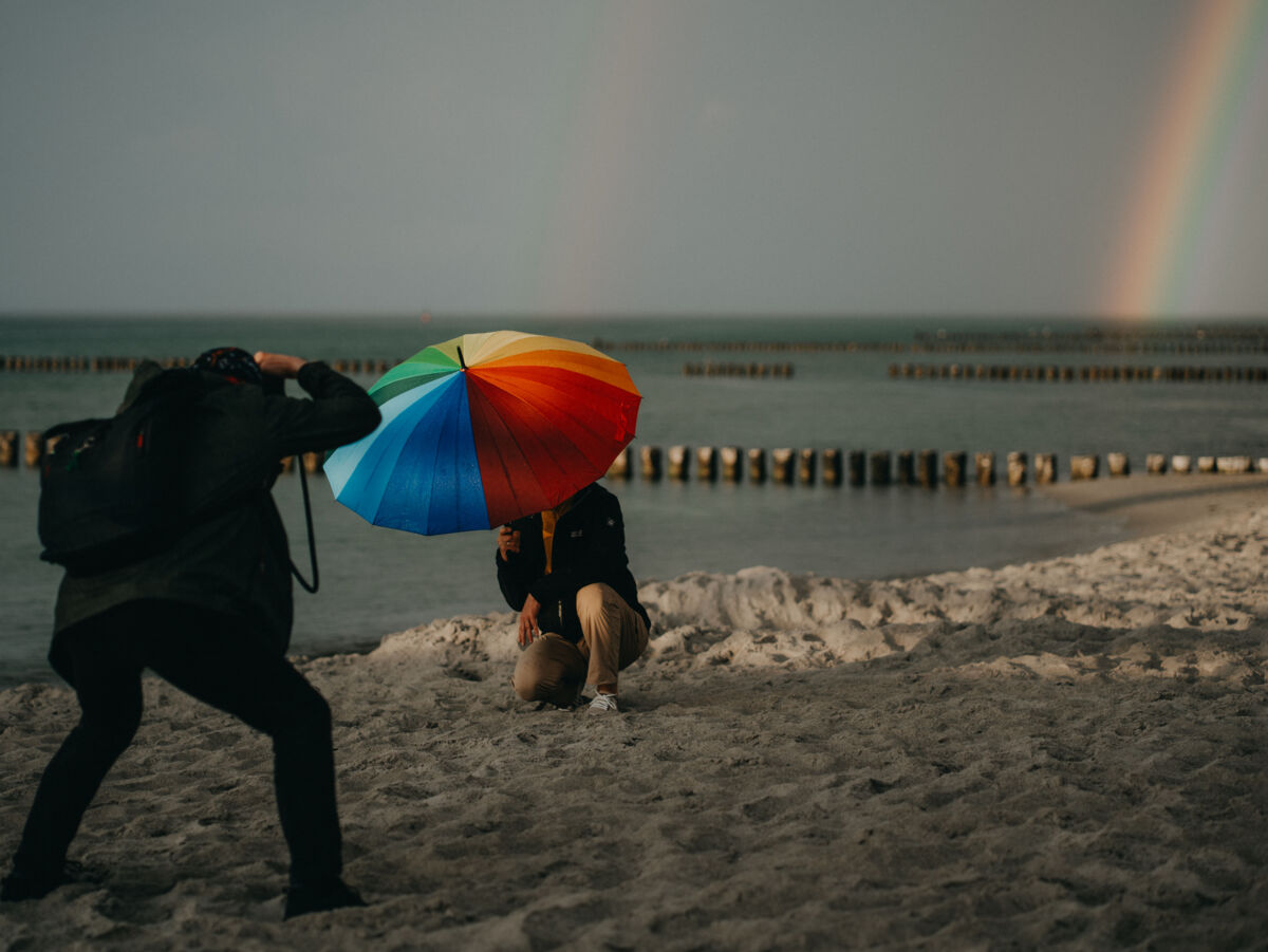 Eine Person fotografiert am Strand eine zweite Person mit einem Schirm in Regenbogenfarben. Im Hintergrund ist ein Regenbogen über dem Meer.