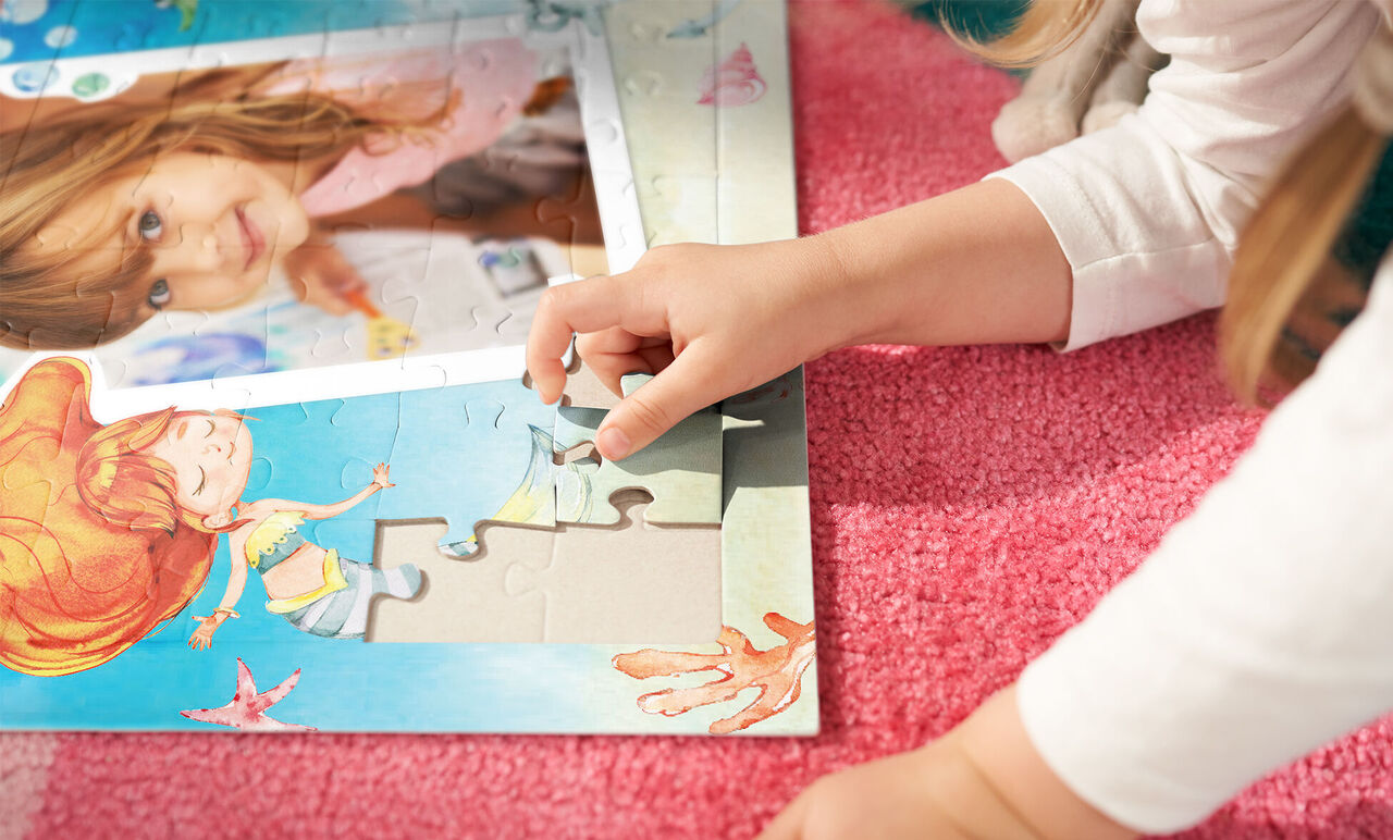 Ein Kind liegt auf einem weichen, pinken Teppich und fügt mit einer Hand ein Puzzlestück in ein buntes Puzzle ein. Im Puzzle sind fröhliche Meereswesen und ein Mädchen abgebildet, das mit ihnen spielt.