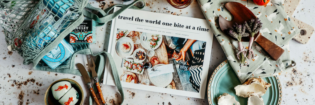 Offenes CEWE FOTOBUCH mit Essensfotos, Text: 'Travel the world one bite at a time', umgeben von Tellern, Muscheln und Tomaten