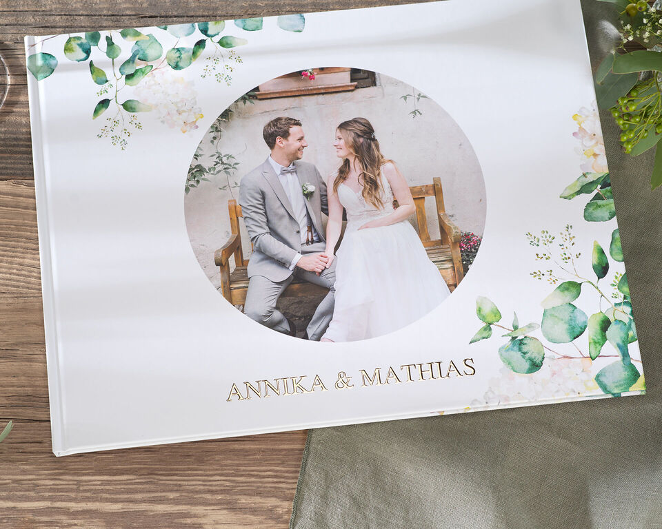 Ein wunderschön gestaltetes CEWE Fotobuch mit einem romantischen Cover. Das Cover zeigt ein rundes Bild eines Paares, umgeben von floralen Elementen und dem Schriftzug „ANNIKA & MATHIAS“.
