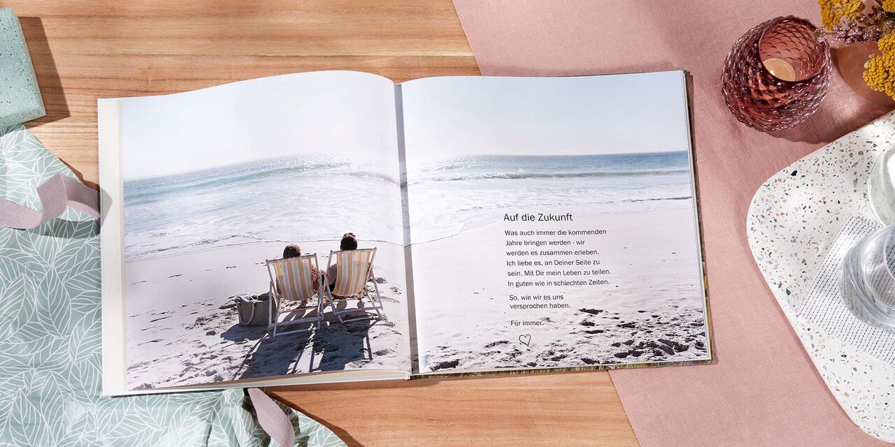 Zwei Personen auf Liegen am Strand, entspannen und genießen den Ausblick auf das Meer, mit einem geöffneten Fotobuch auf der Seite.