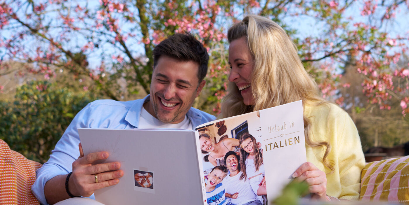 Zwei Personen sitzen draußen und blättern gemeinsam in einem Fotobuch mit Familienbildern.