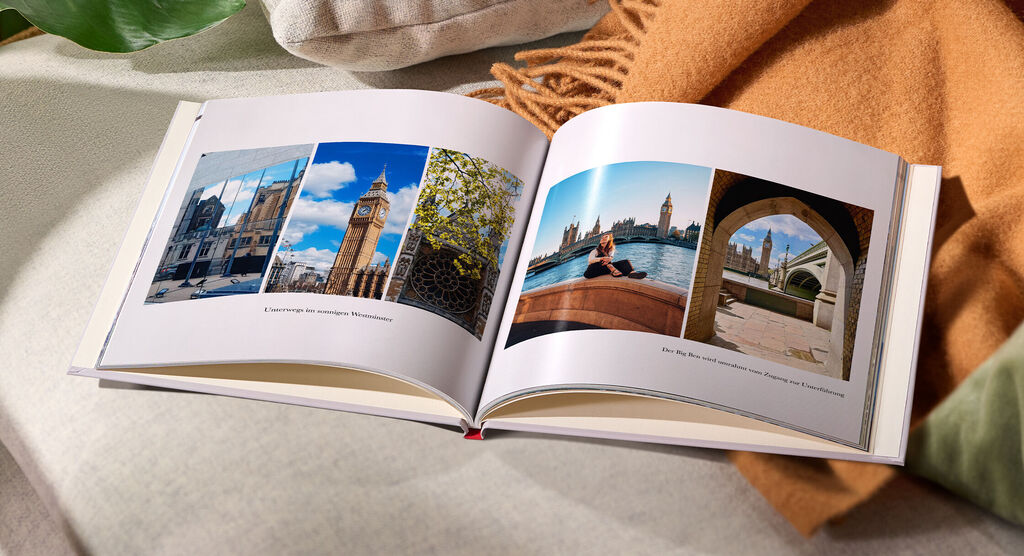 Ein geöffnetes Fotobuch mit bunten Urlaubsbildern, das auf einem gemütlichen Sofa liegt, umgeben von kuscheligen Textilien.