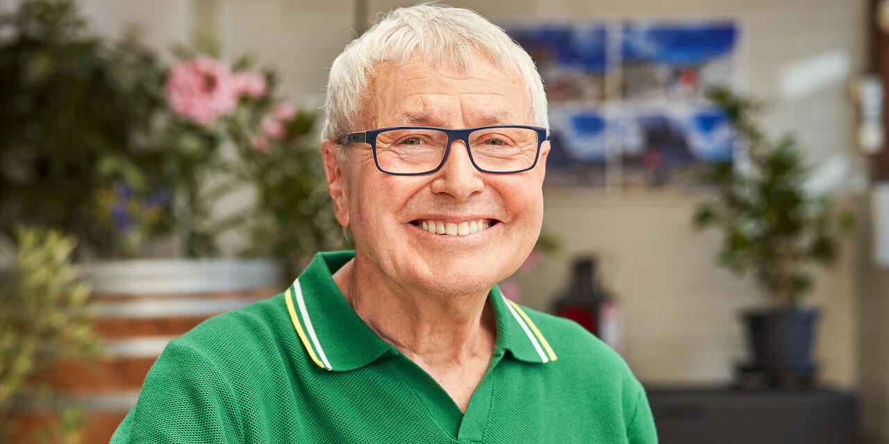 Ein älterer Herr in einem grünen Polohemd lächelt vor einer Blüten- und Pflanzenkulisse.