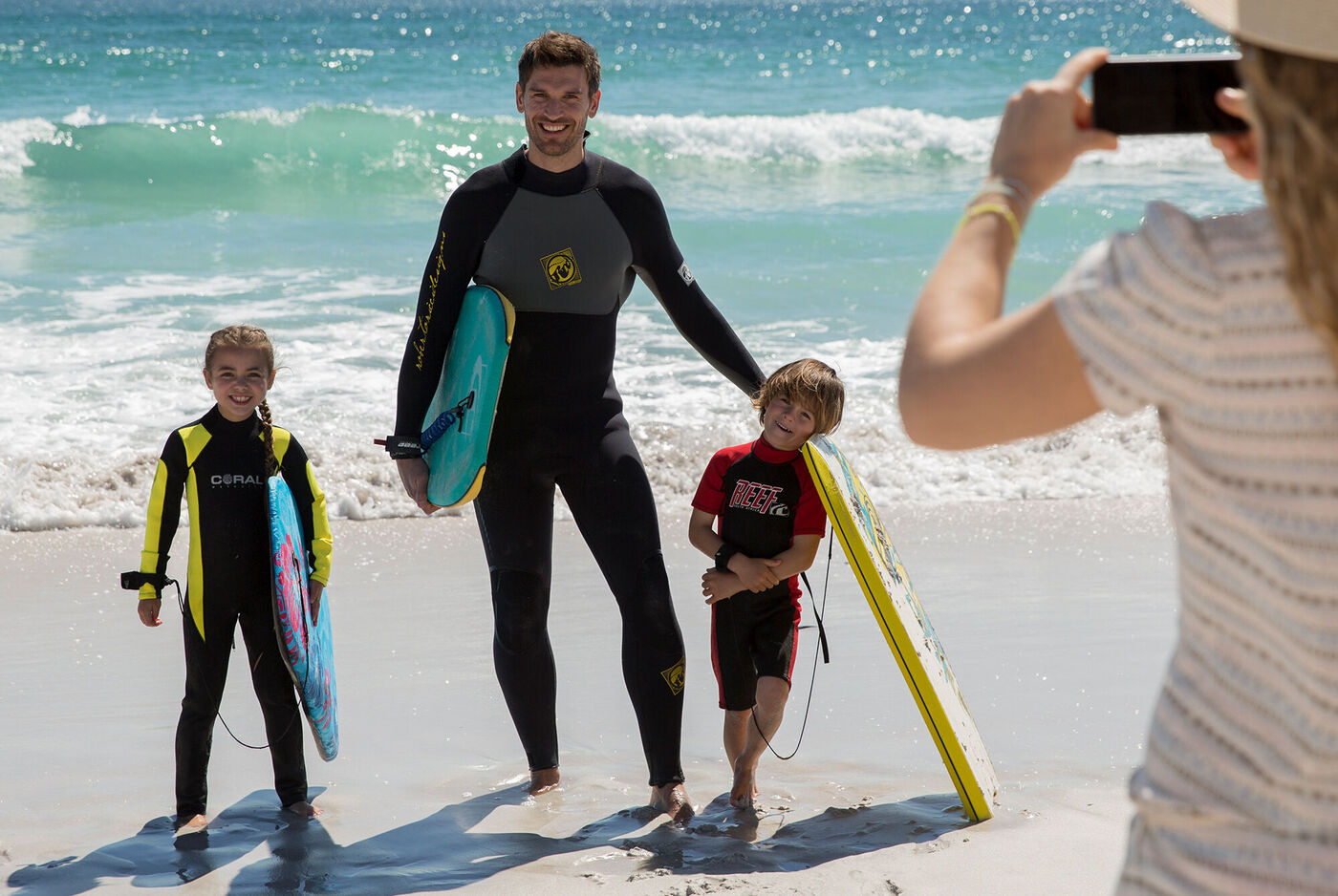 Ein Vater steht mit zwei Kindern am Strand, alle mit Surfbrettern, während ein Foto gemacht wird