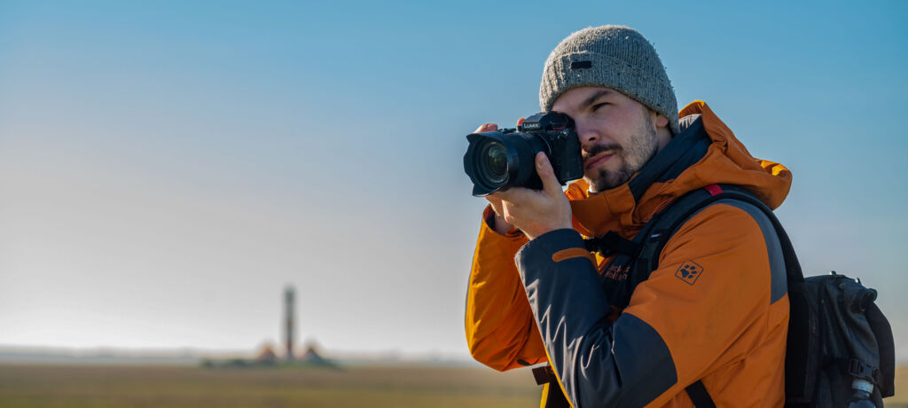 Ein Mann in oranger Winterjacke und mit Mütze steht vor einer flachen Landschaft mit blauem Himmel. In der Hand hält er eine Kamera.
