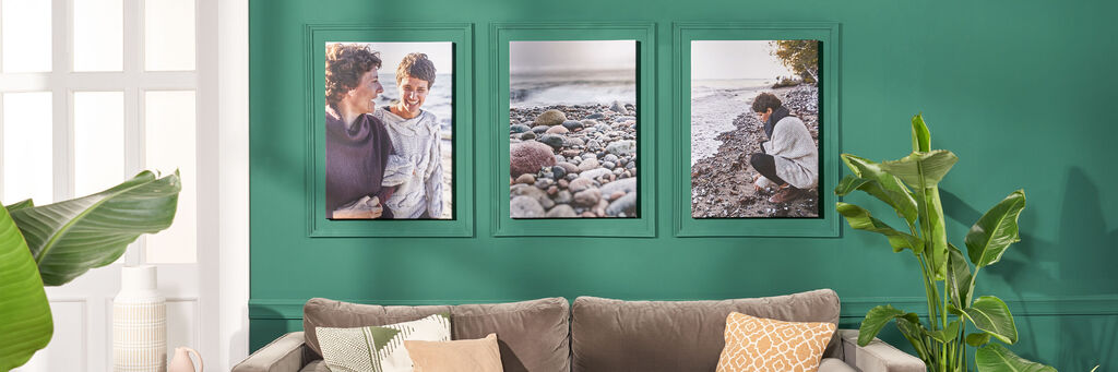 Drei Bilder an einer grünen Wand, die besondere Momente zeigen. Eines zeigt eine Frau und ein Kind. Das zweite Bild zeigt eine Frau, die Steine am Strand betrachtet. Das dritte Bild zeigt eine beruhigende Landschaft mit Wasser.