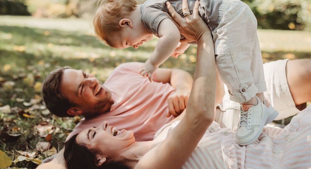 Eine liebevolle Familie genießt Zeit im Freien. Ein Elternteil hält das kleine Kind hoch, während die andere Person entspannt im Gras liegt.