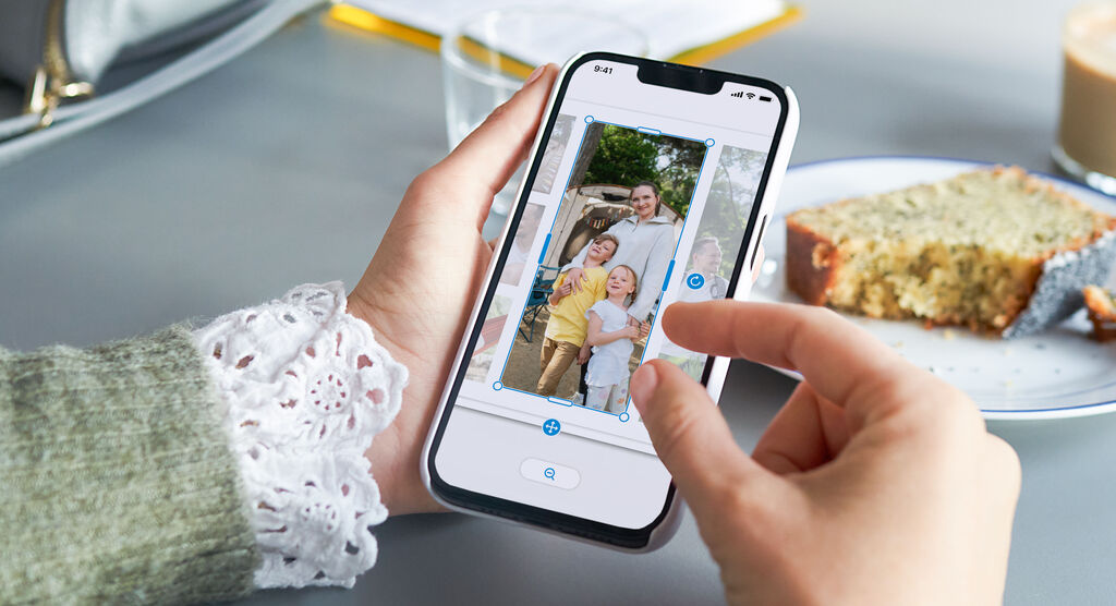 Person hält Smartphone, auf dem in der CEWE App ein Familienfoto zugeschnitten und ausgerichtet wird; Kuchen daneben.