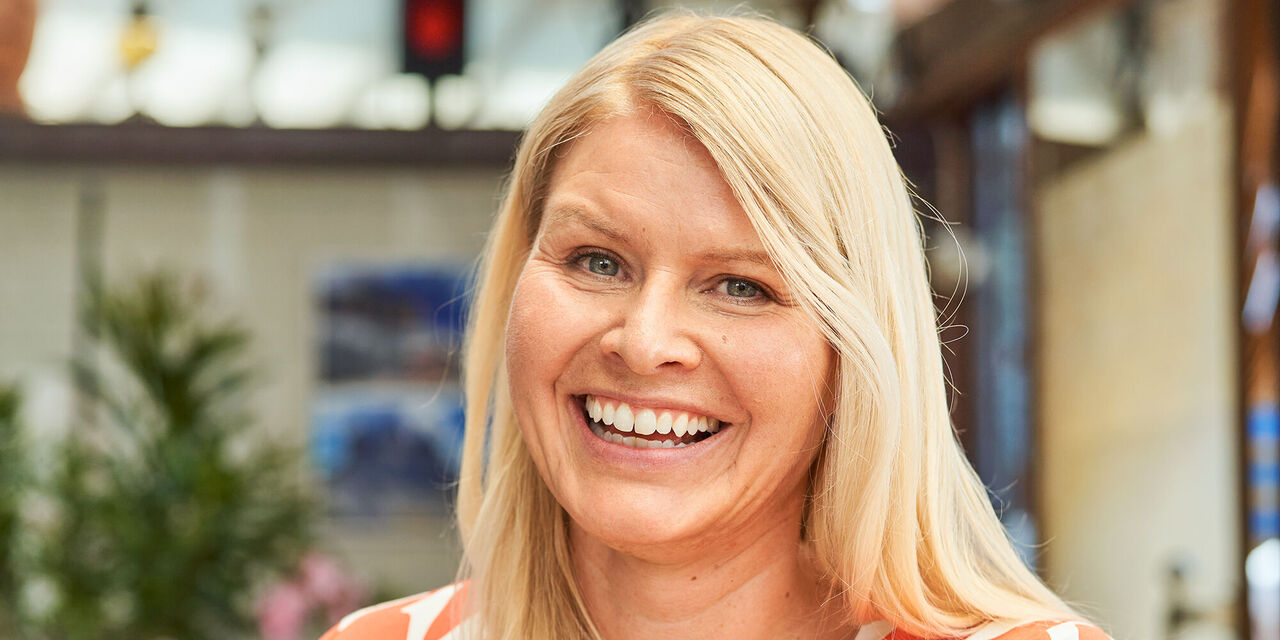 Eine Frau mit blonden Haaren in einer blühenden Umgebung, lächelt voller Freude.