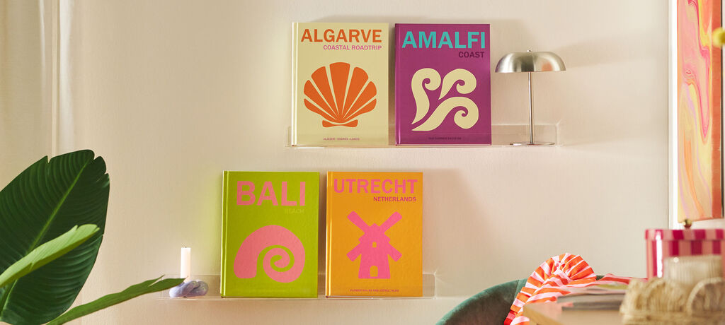 In einem Wohnzimmer hängen zwei Bilderleisten an der Wand, auf denen jeweils zwei Reisebücher in bunten Farben platziert sind.