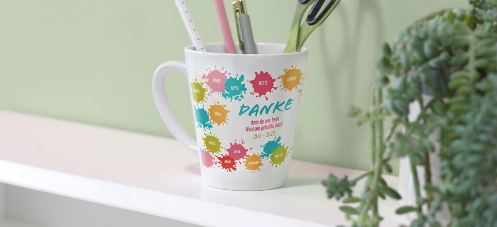 Eine Tasse mit bunten Farbklecksen und dem text "DANKE" steht auf einem Tisch.