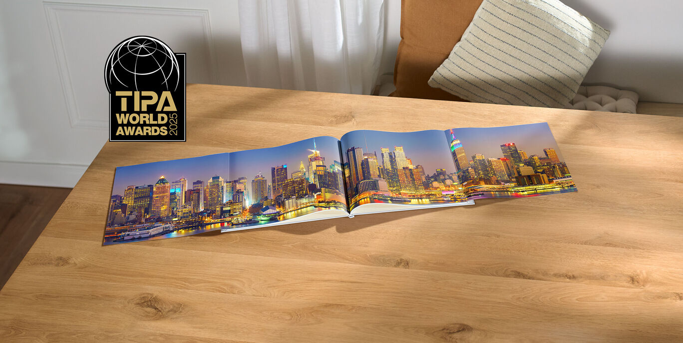 Ein offenes CEWE FOTOBUCH zeigt eine schöne Skyline einer Stadt bei Nacht. Die Lichter der Gebäude funkeln und spiegeln sich im Wasser. Der Tisch ist aus hellem Holz und eine Stuhlecke ist im Hintergrund zu sehen.