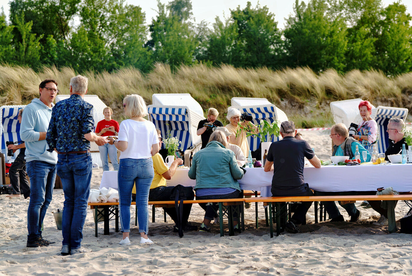 Mehrere Menschen sitzen an einem langen Tisch am Strand und unterhalten sich.