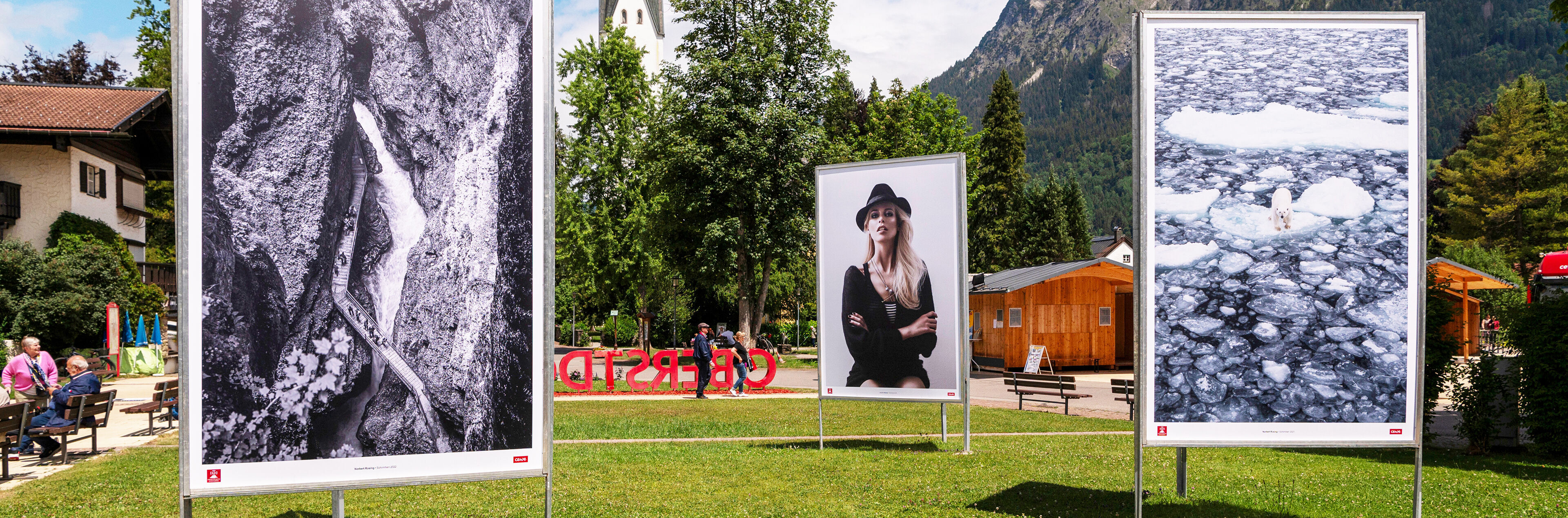 Auf einer Wiese stehen drei große Fotografien. Im Hintergrund ist eine Kirche zu sehen.