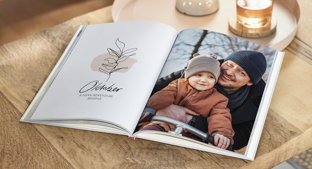 Ein geöffnetes Fotobuch auf einem Tisch zeigt eine romantische Illustration und ein Bild von einem Elternteil, der mit einem Kind spielt.