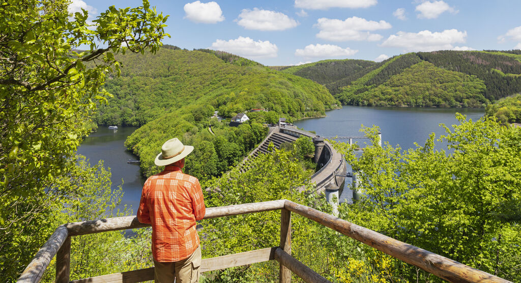 Mann in orange kariertem Hemd mit Hut steht an Aussichtspunkt und schaut auf den See umgeben von grüner Natur