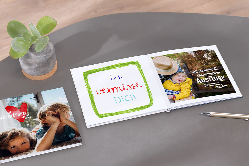 Ein offenes CEWE FOTOBUCH auf einem Tisch. Es zeigt handgeschriebene Botschaften und Fotos von Kindern und Ausflügen. Eine Pflanze steht daneben, die eine fröhliche Atmosphäre schafft.