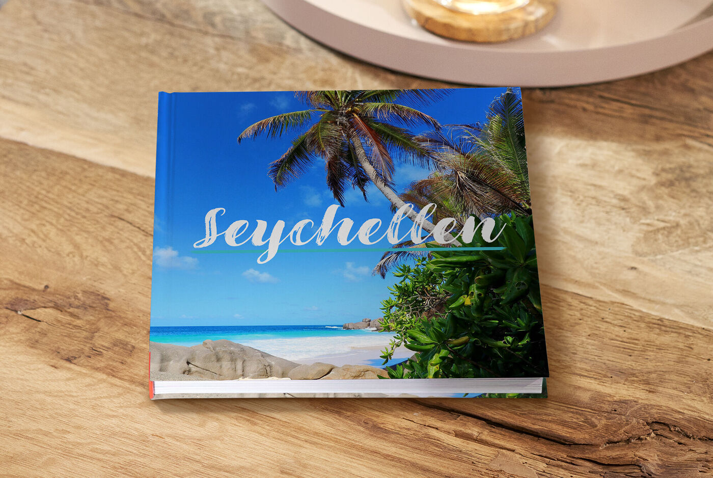 Ein wunderschönes CEWE FOTOBUCH mit dem Titel 'Seychellen' auf einem Holztisch, umgeben von grünen Pflanzen und blauer Himmel.
