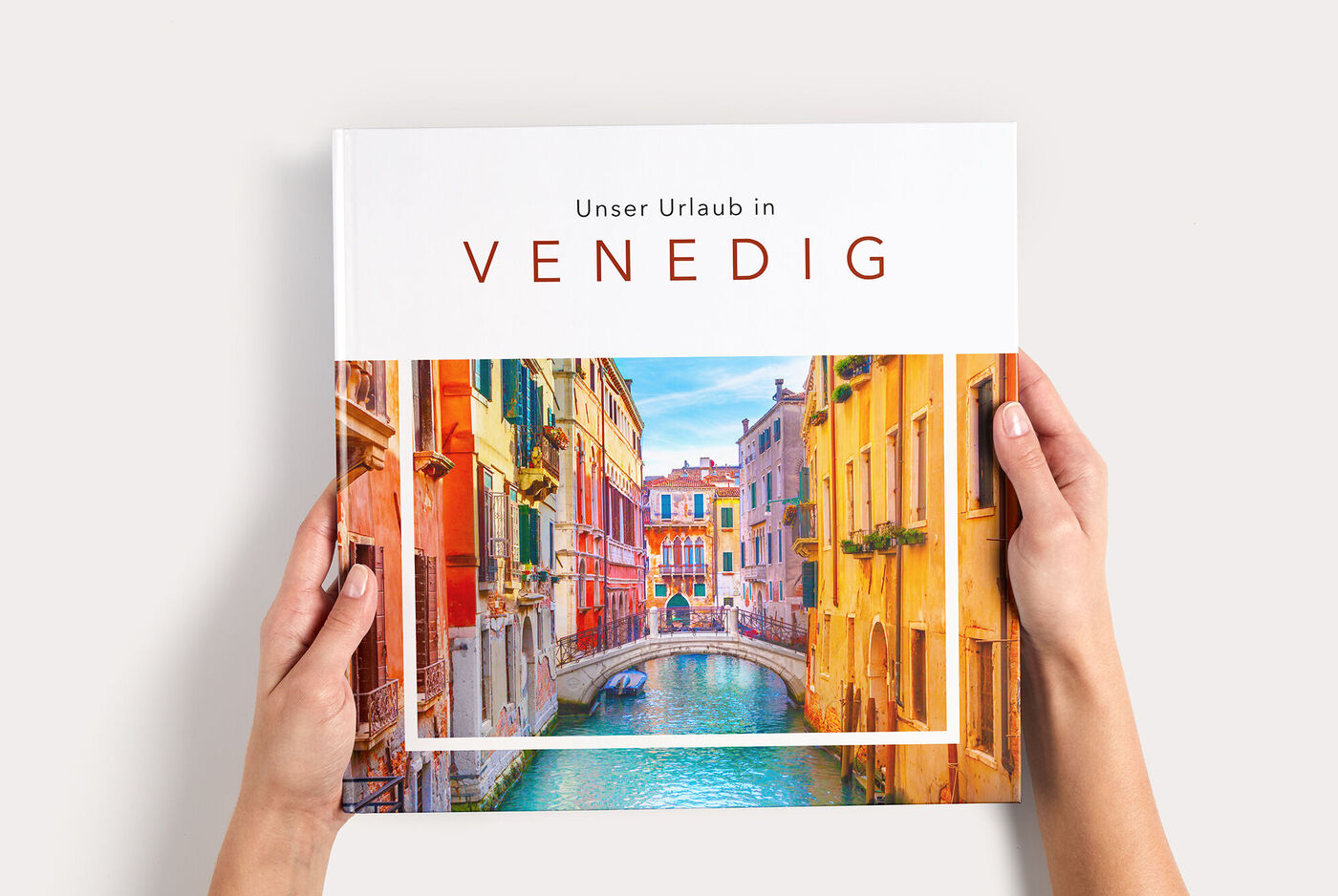 Hände halten ein CEWE FOTOBUCH XL mit bunten venezianischen Häusern am Kanal und Schriftzug "VENEDIG"