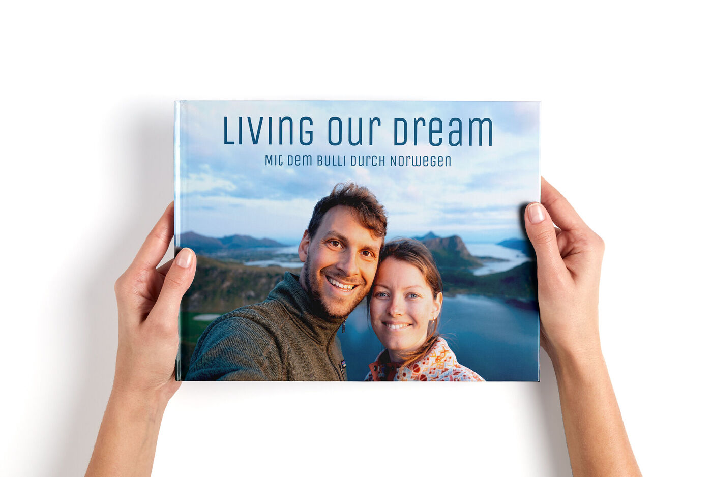 Zwei Personen halten ein CEWE FOTOBUCH mit dem Titel 'Living Our Dream' vor einer malerischen Berglandschaft.