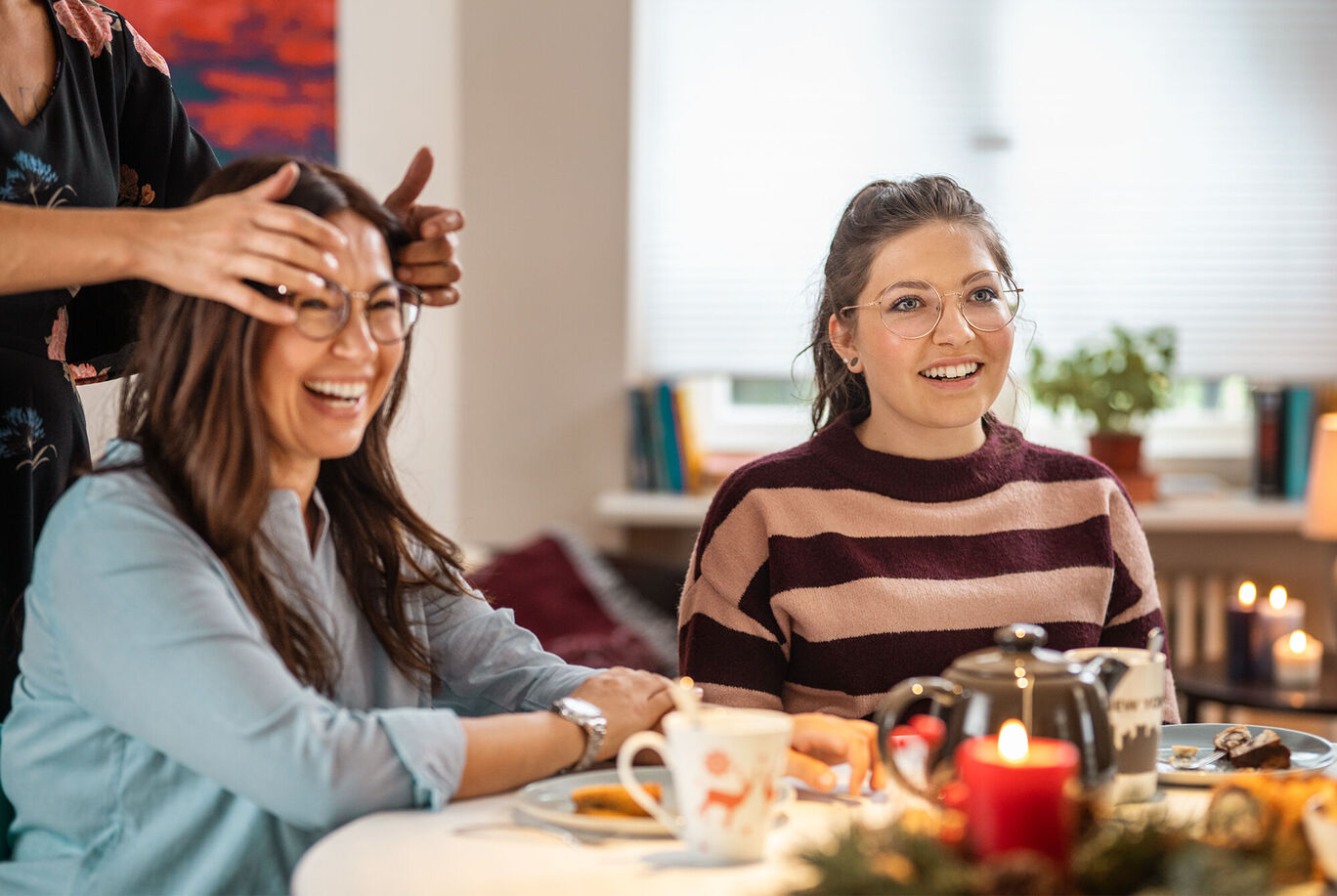 Zwei Personen sitzen an einem Tisch mit Getränken und Snacks, eine Person frisiert die Haare der anderen.