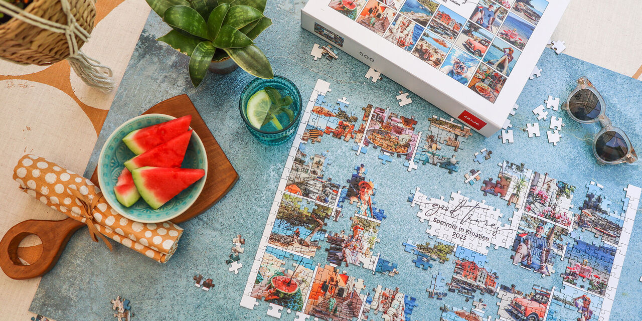 Ein buntes Puzzle mit Motiven von Urlaubsshots liegt auf einem blauen Tisch, umgeben von saftiger Wassermelone und einem Glas Limonade