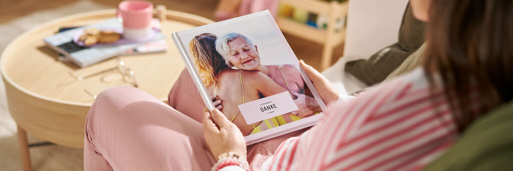 Eine Frau sitzt auf einem Sofa im Wohnzimmer, in ihren Händen hält sie ein CEWE FOTOBUCH mit der Aufschrift „Danke“.