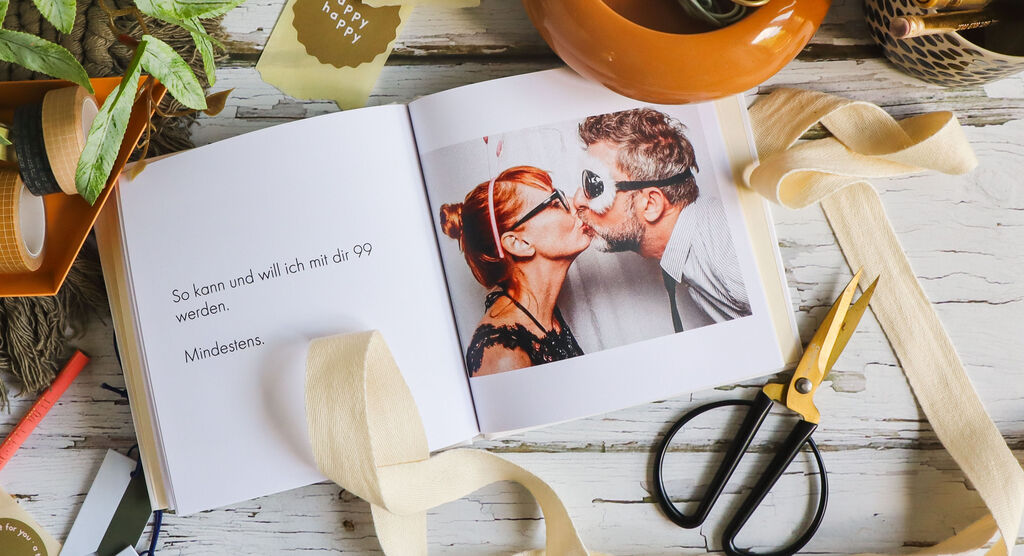 Ein geöffnetes Fotobuch mit einer liebevollen Aufnahme eines Paares, umgeben von Deko-Elementen wie einer Schere und Naturmaterialien