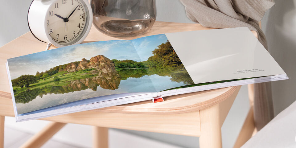 Auf einem Holztisch liegt ein aufgeklapptes CEWE Fotobuch mit Bildern einer Landschaft, die sanften Hügel und einen See zeigt. Eine Uhr und eine Glasvase dekorieren den Tisch.