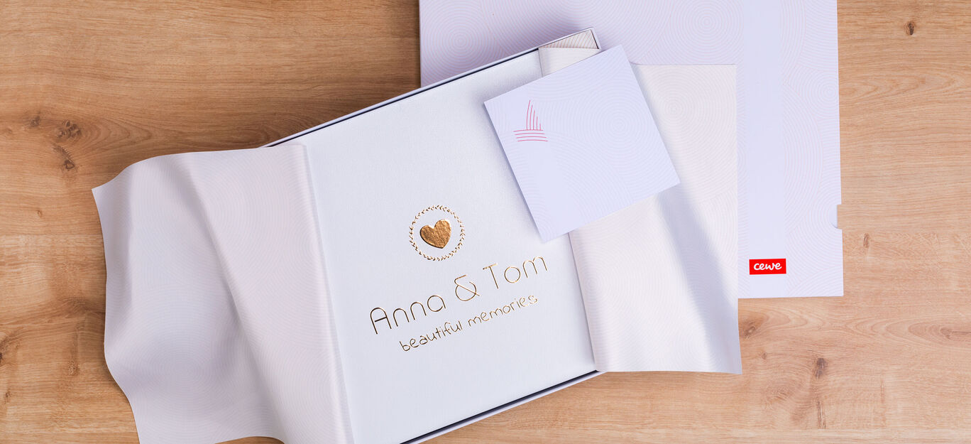 Ein geöffnetes CEWE FOTOBUCH liegt auf einem Holztisch. Es zeigt elegante Gestaltung mit Herz, Namen und einer Grußkarte.