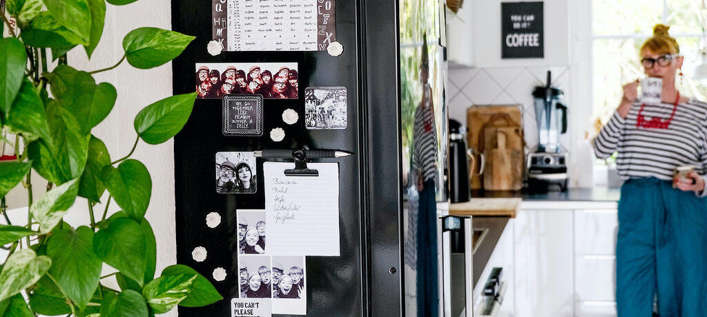 Ein Kühlschrank mit Erinnerungsfotos und Notizen. Im Hintergrund steht eine Person und hält ein Glas Wasser.