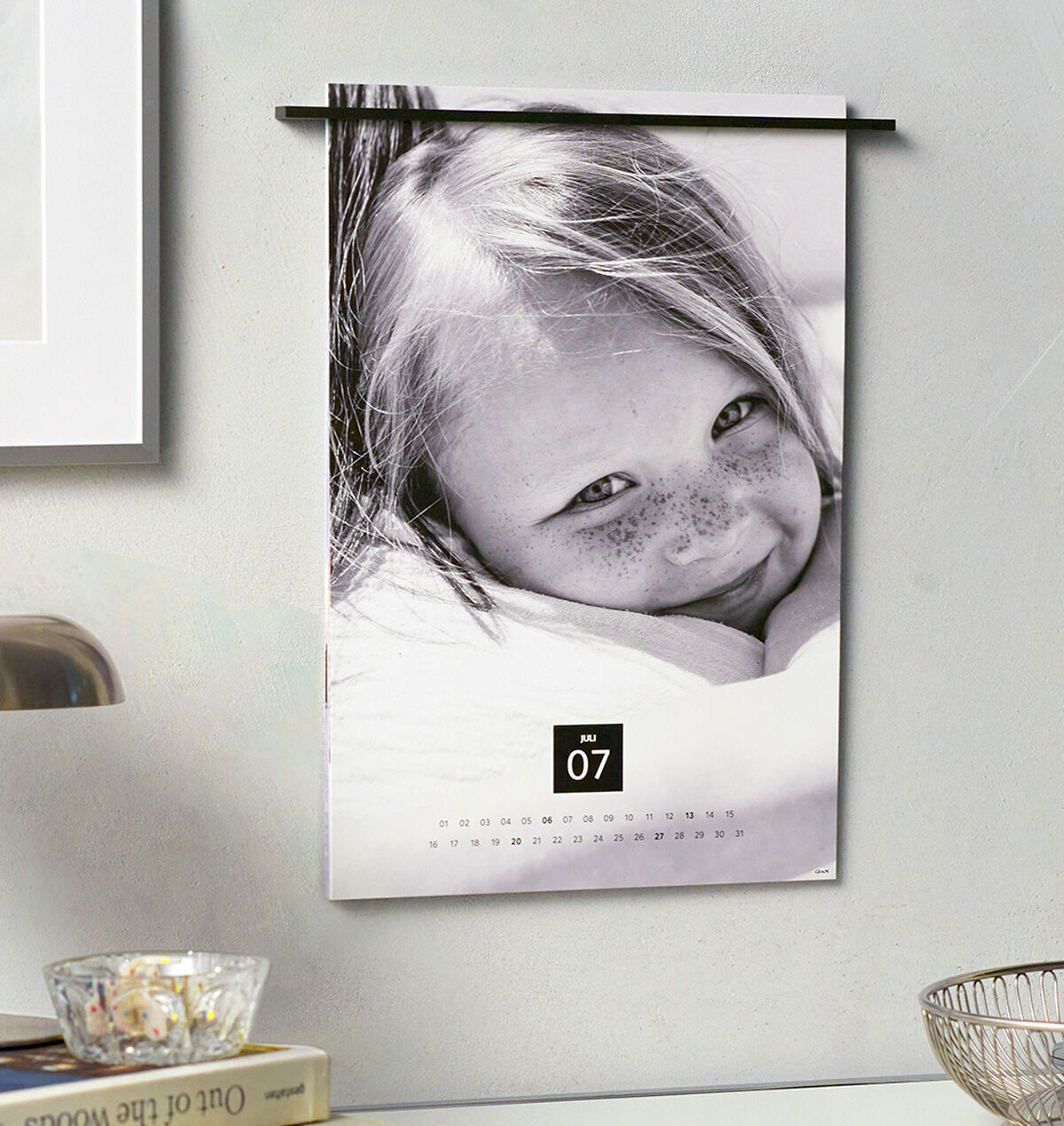Schwarz-weißer CEWE Fotokalender hängt an einer hellen Wand über einem Schreibtisch mit Lampe, Deko-Schale und Büchern.",