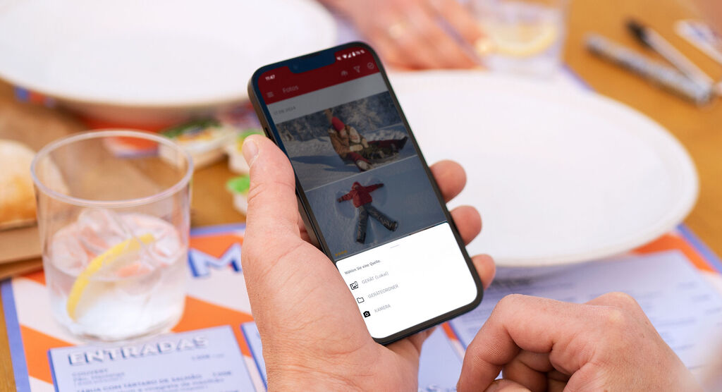 Eine Hand hält ein Smartphone, auf dem Urlaubsfotos angezeigt werden, inklusive eines Schlittenfahrers im Schnee und eines Schneeengels