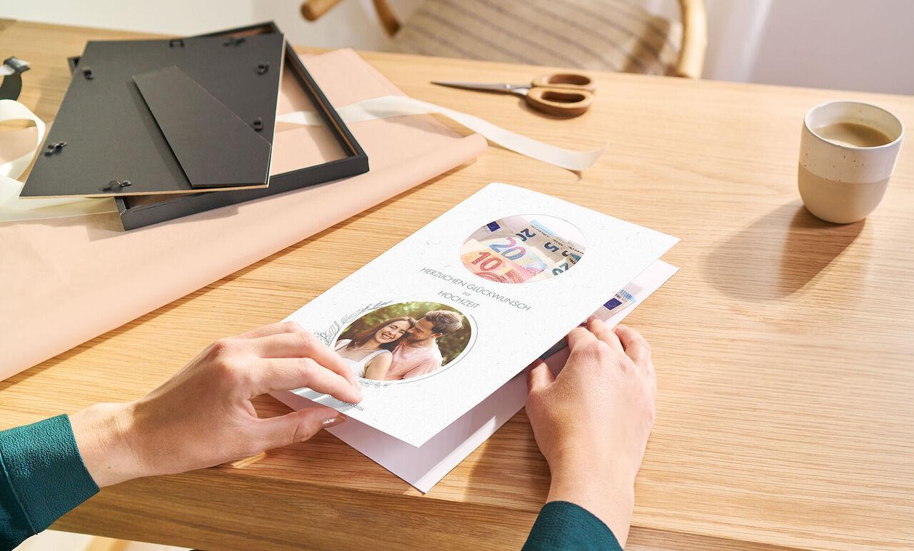 Händed eines Menschen halten eine personalisierte Gratulationskarte mit Fotos an einem hellen Holztisch.