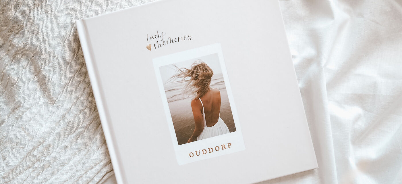 Eine Frau mit langem Haar in einem weißen Kleid von hinten am Strand, auf dem Cover eines CEWE Fotobuchs mit dem Titel "OUDORP".