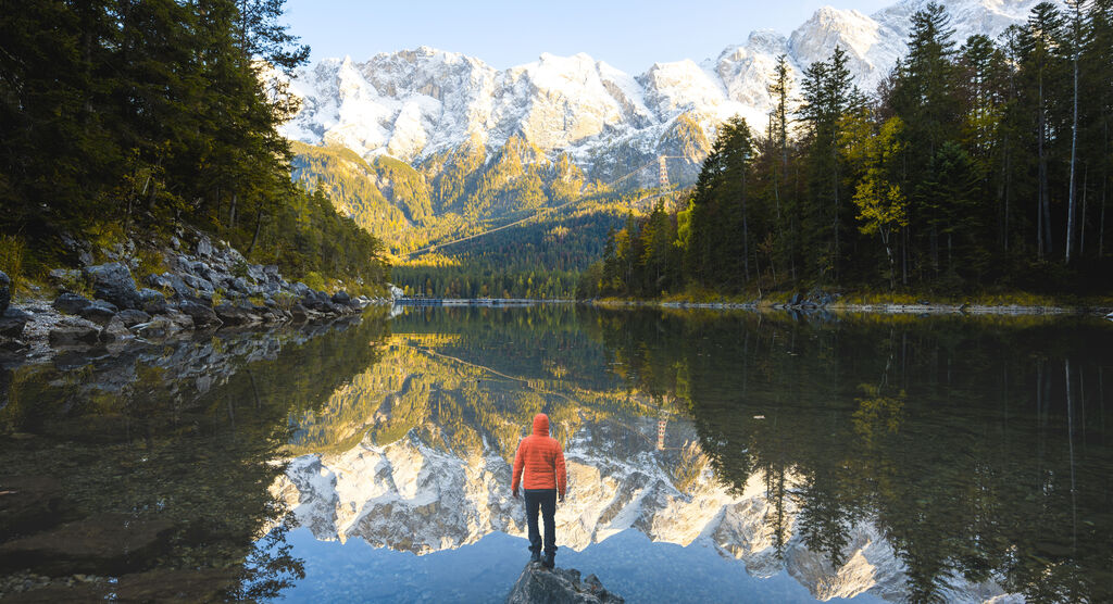 Ein Mensch in orangefarbener Jacke steht an einem ruhigen See, umgeben von Bergen und Bäumen. Die Bergspitzen sind mit Schnee bedeckt und spiegeln sich im klaren Wasser.