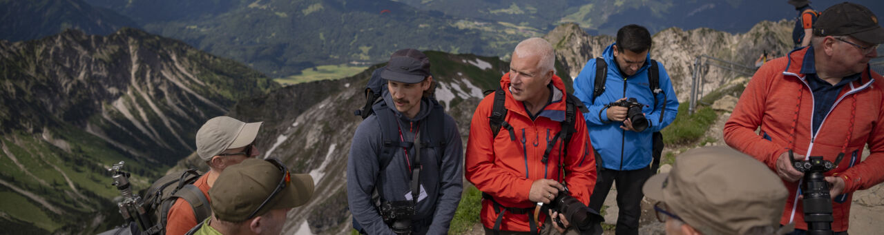 Mehrere Personen in Outdoor-Outfits und mit Kameras in den Händen stehen diskutierend auf einem Berggipfel.