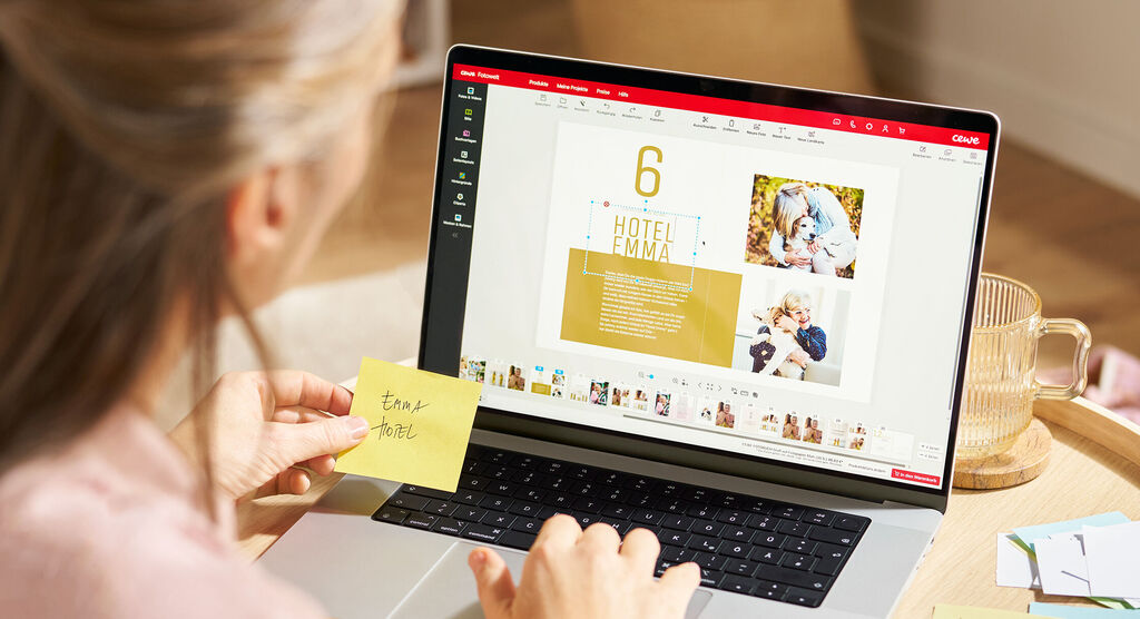 Eine Frau sitzt im Wohnzimmer an einem Couchtisch, auf dem ein Laptop steht. In der Hand hält sie einen beschrifteten Klebezettel.