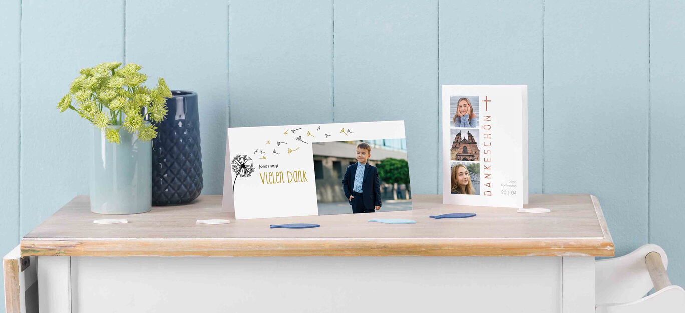 Zwei CEWE Grußkarten und ein CEWE Fotobuch auf einem Tisch mit einer Pflanze im Hintergrund