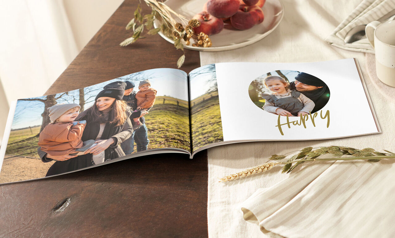 Ein offenes Fotobuch auf einem Tisch mit einer fröhlichen Familie im Freien. Auf einer Seite steht das Wort 'Happy' in goldener Schrift.