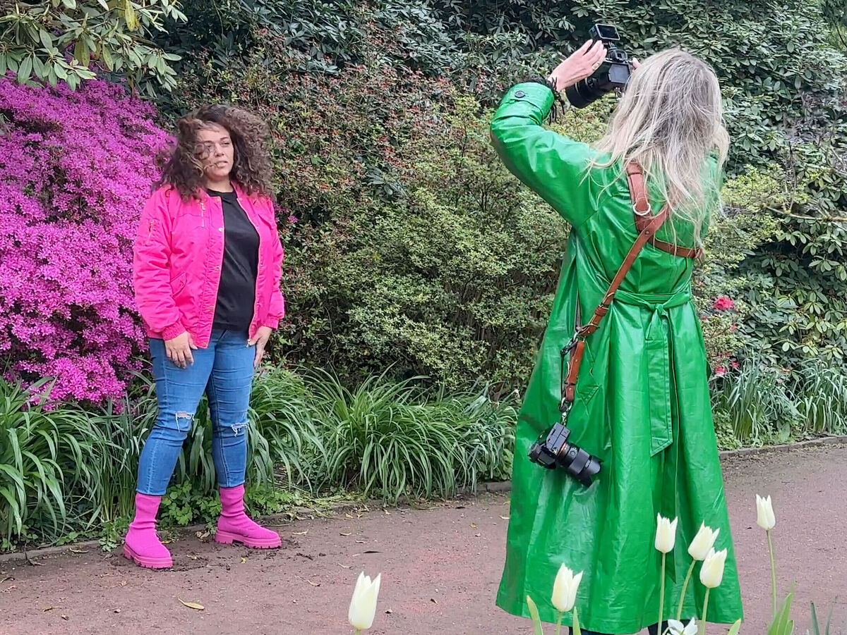 Eine Person in einem pinken Outfit posiert vor bunten Blumen, während eine zweite Person in einem grünen Mantel fotografiert