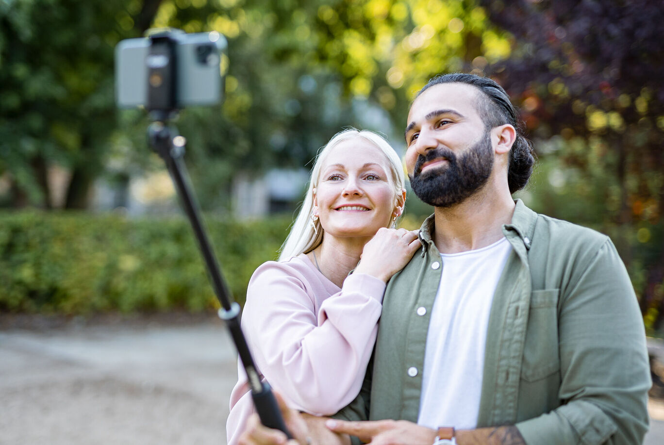 Ein Paar macht ein Selfie im Park mit einem Smartphone. Die Umgebung ist grün und sonnig.