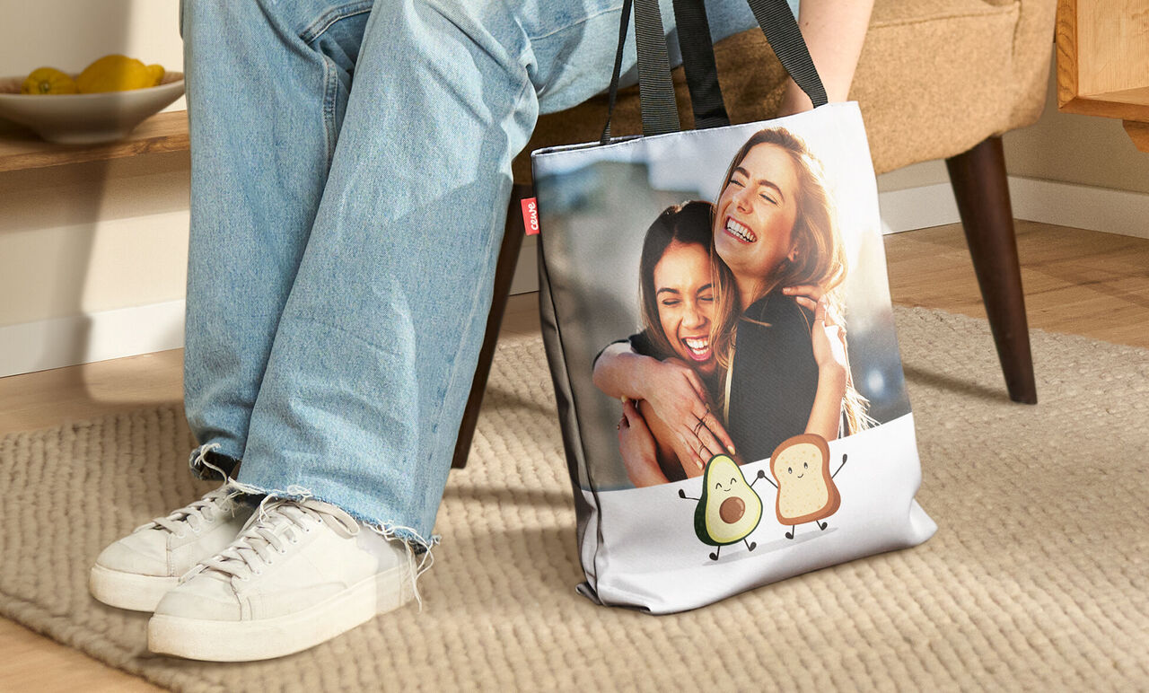 Eine Person sitzt auf einem beigen Teppich mit einer personalisierten Tasche mit Avocado-Motiven. Im Hintergrund ist ein Sofa und ein Obstkorb sichtbar