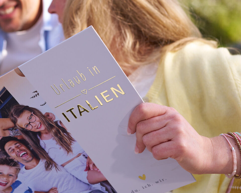 Person hält ein Fotobuch mit der goldenen Aufschrift „Urlaub in Italien“.