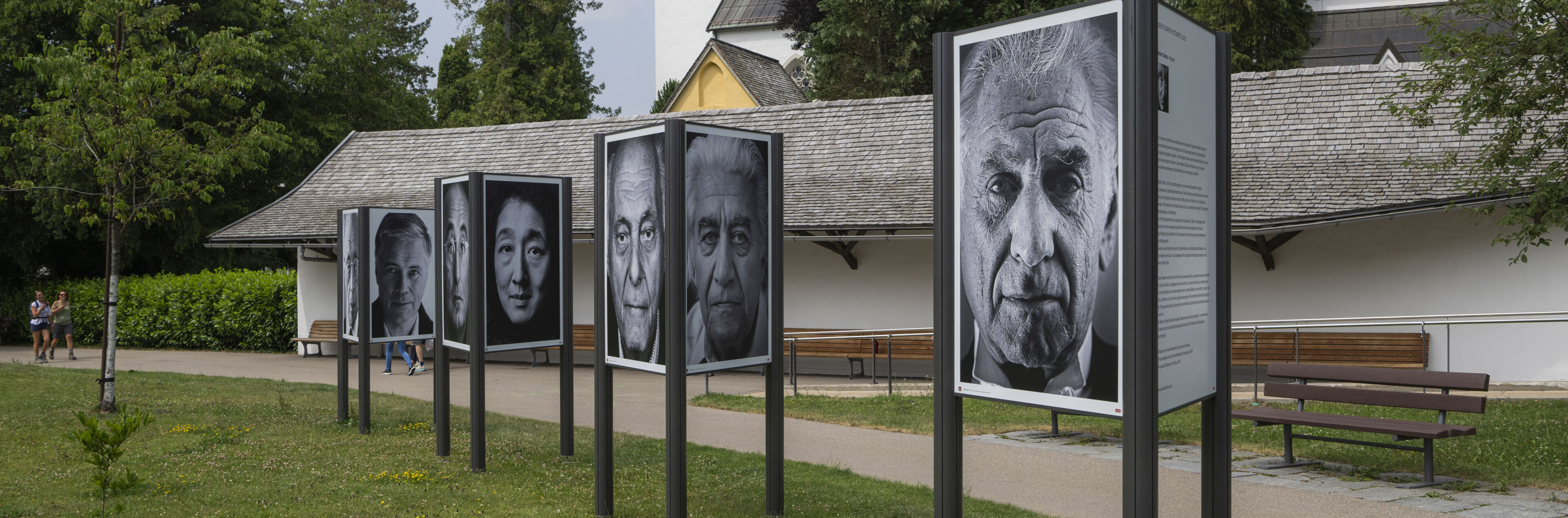 Drei große Aufsteller auf einer Wiese zeigen verschiedene Schwarz-Weiß-Porträts. Im Hintergrund ist ein Kirchturm.