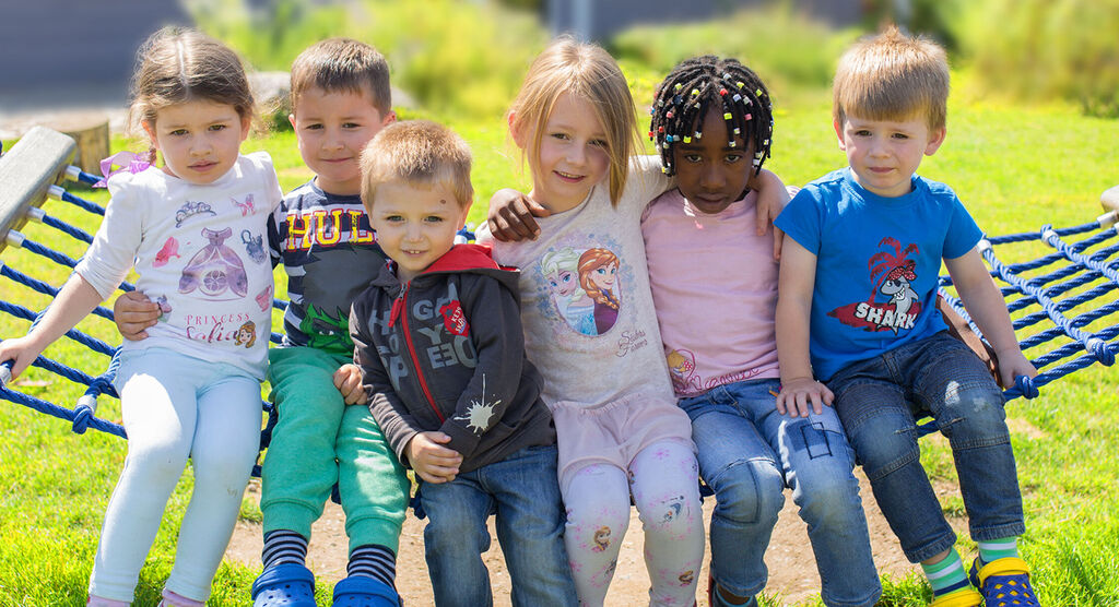 Sechs Kinder sitzen fröhlich auf einer Schaukel im Freien. Sie tragen bunte Kleidung und genießen den sonnigen Tag.